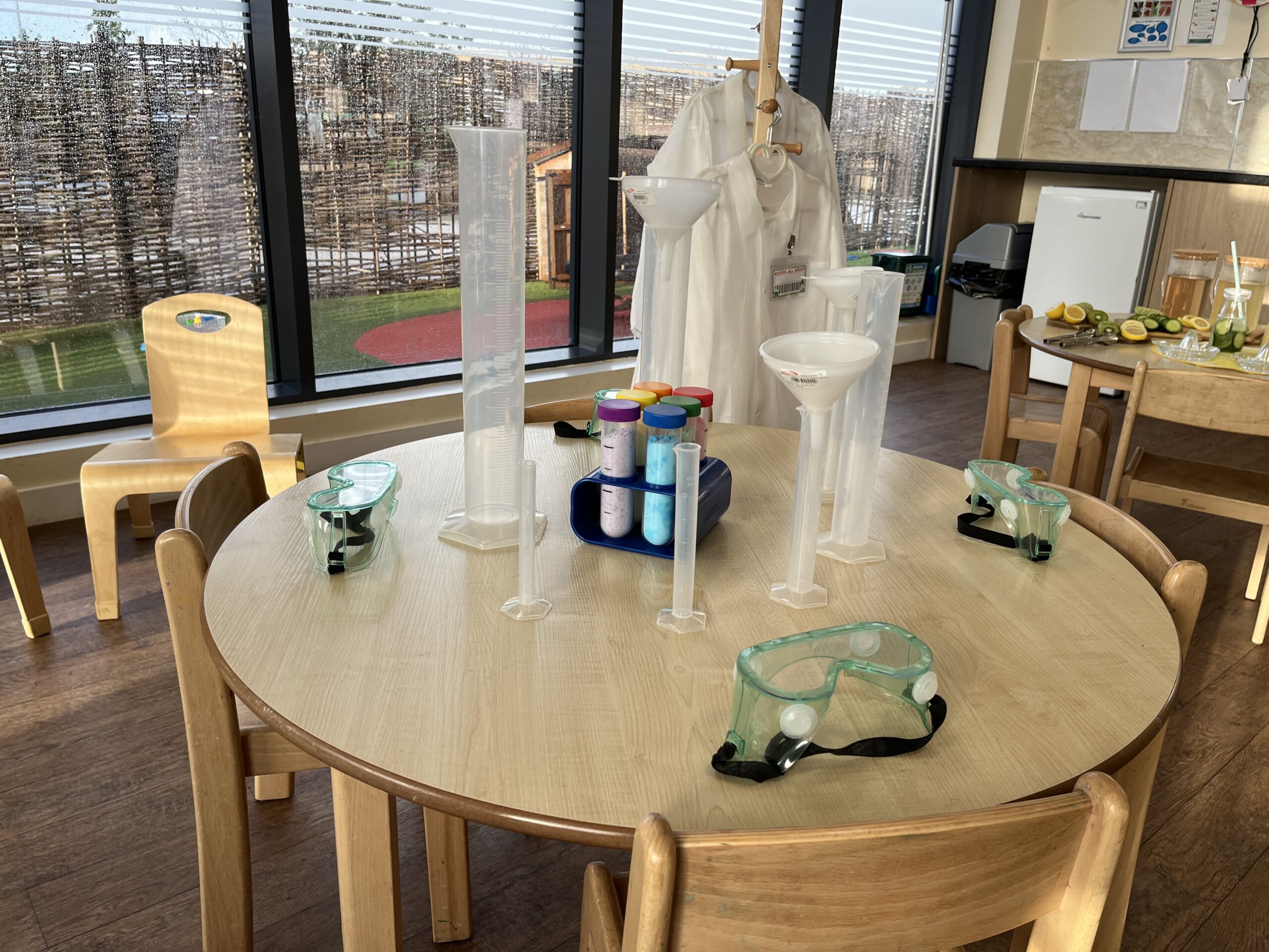 a stem activity set up on one of the tables in pre school including lots of different mixing and measuring jugs, with childrens lab coats hung up to the right