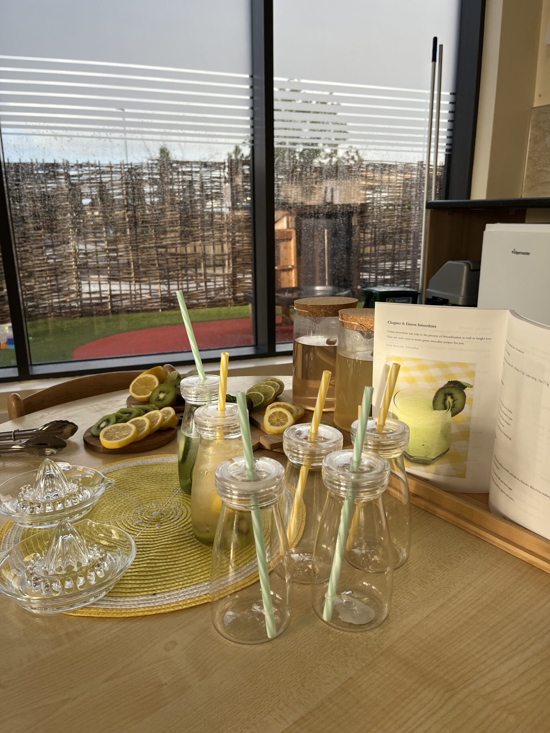 a table with cups, slices of kiwi and lemon, jugs and a recipe book for how to make a drink