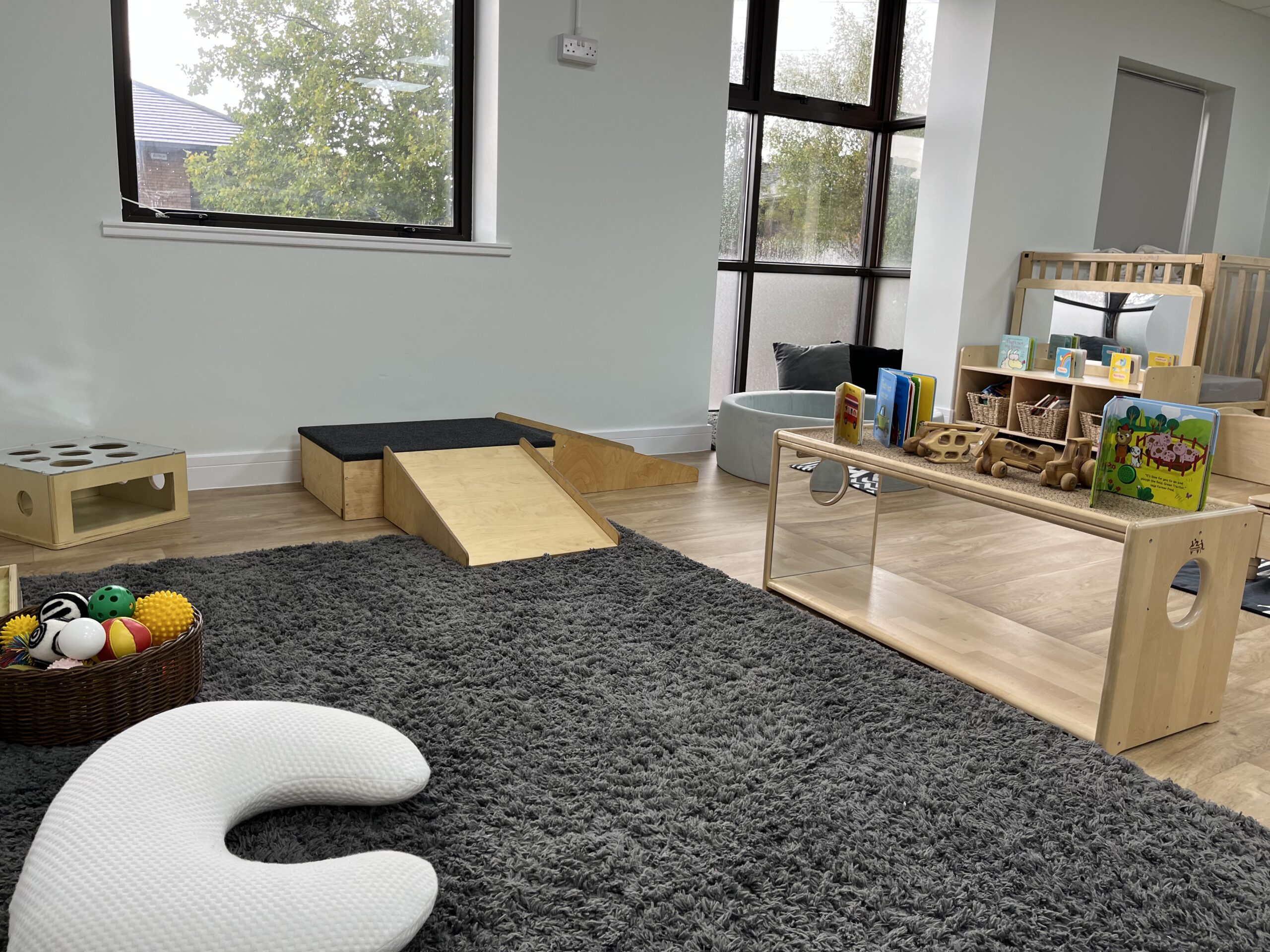 Baby room , showing baby support pillow, baby crawling ramp, basket of toys and mirrored shelf with books and wooden toys on