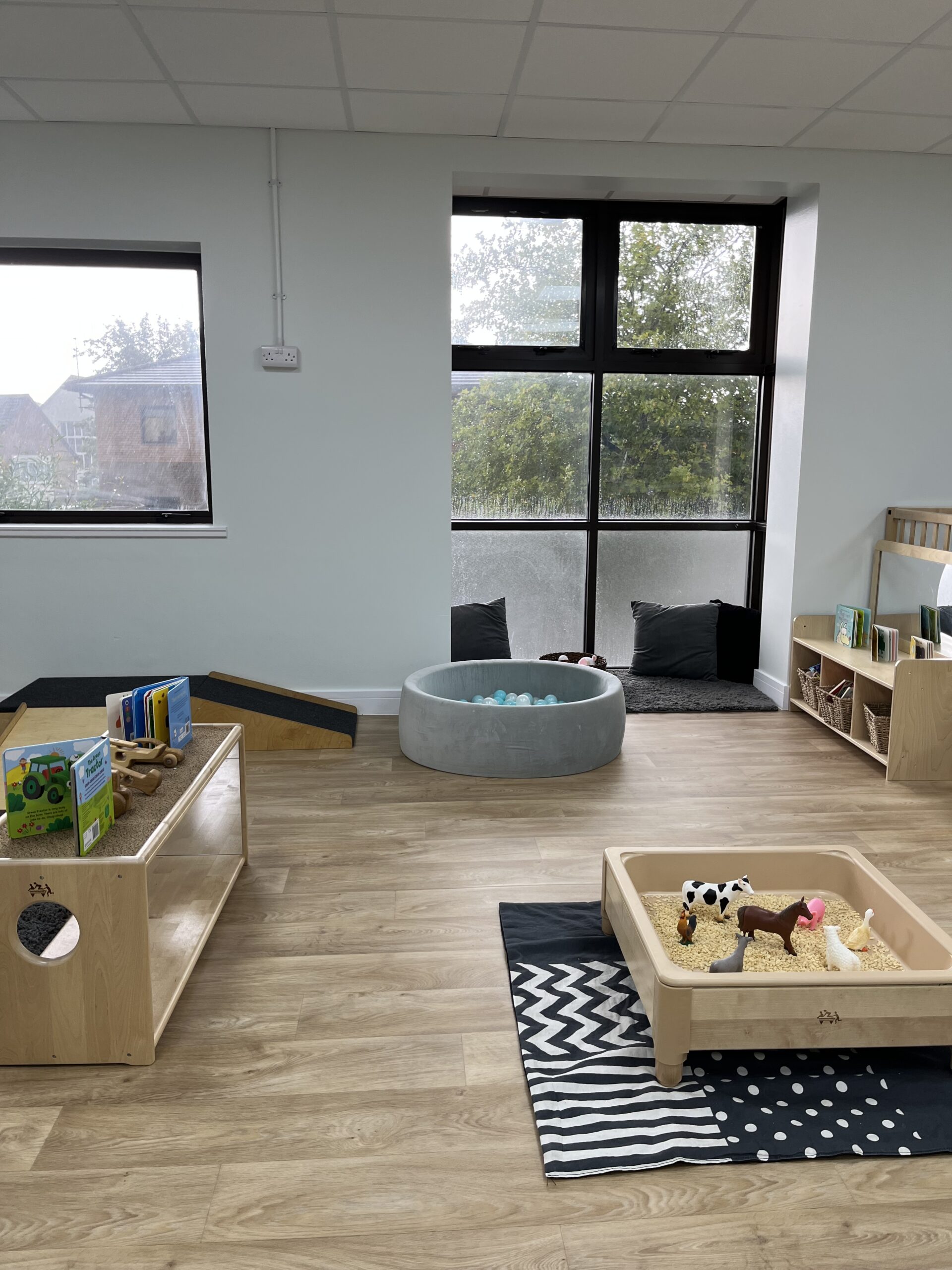 Baby room corner showing a ball pit, farm animal themed tuff tray and multiple mirrored tables with books and wooden cars placed ontop.